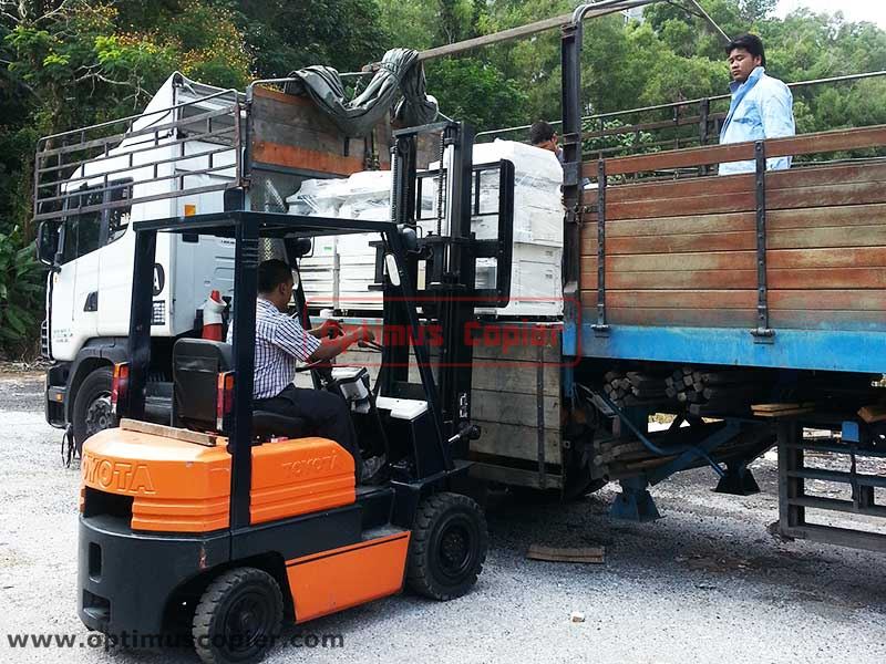 Optimus Copier's staff operating an orange forklift is loading stacked copier machines onto a lorry, demonstrating copier rental and delivery service across Malaysia especially Klang Valley, Melaka and Seremban.
